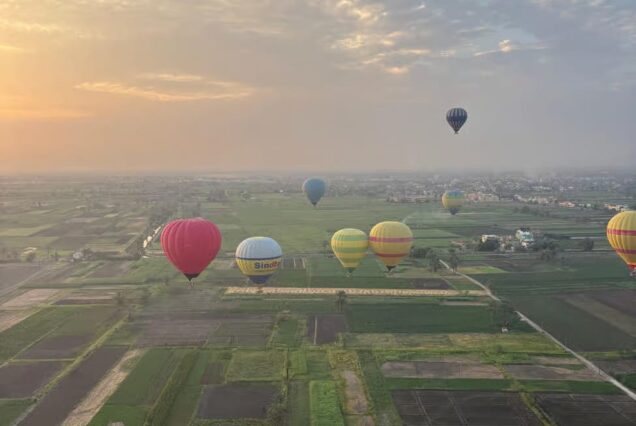 Hot air balloons floating at sunrise over Luxor, Egypt, with fields and ancient landscapes visible below.