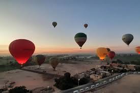 Hot air balloon flying over Luxor, Egypt at sunrise, with the Nile River and West Bank temples visible below.
