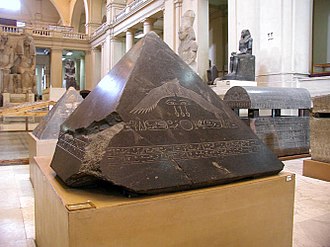 The pyramidion of Amenemhat III, carved from black granite, decorated with a winged sun disk, uraeus cobras, and hieroglyphs.