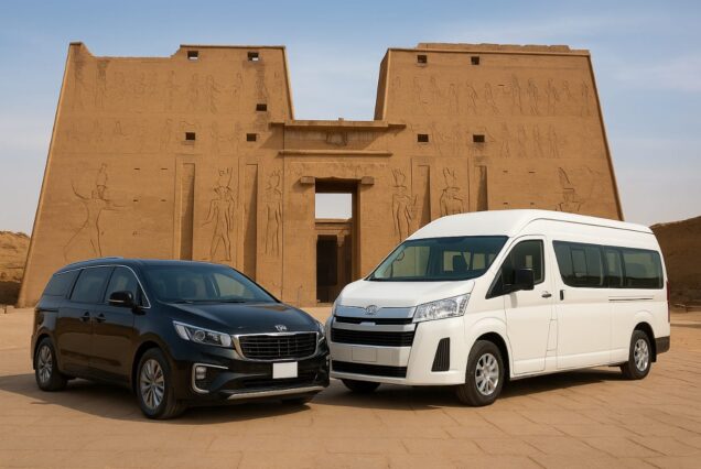 Two vehicles, a black minivan and a white passenger van, parked in front of the Great Temple of Edfu in Egypt.
