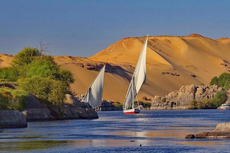 Traditional Egyptian felucca sailboats with large triangular sails gliding on the Nile River in Aswan, surrounded by desert dunes and rocky landscapes.