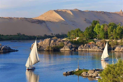 Scenic view of the Nile River in Aswan, Egypt, with felucca sailboats gliding across calm waters surrounded by rocky islands and desert hills.