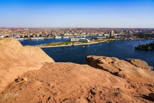 Panoramic view of the Nile River in Aswan, Egypt, with desert landscapes and distant urban areas, taken from an elevated viewpoint.