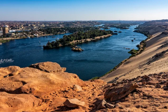 Panoramic view of the Nile River in Aswan, Egypt, with desert landscapes and distant urban areas, taken from an elevated viewpoint.