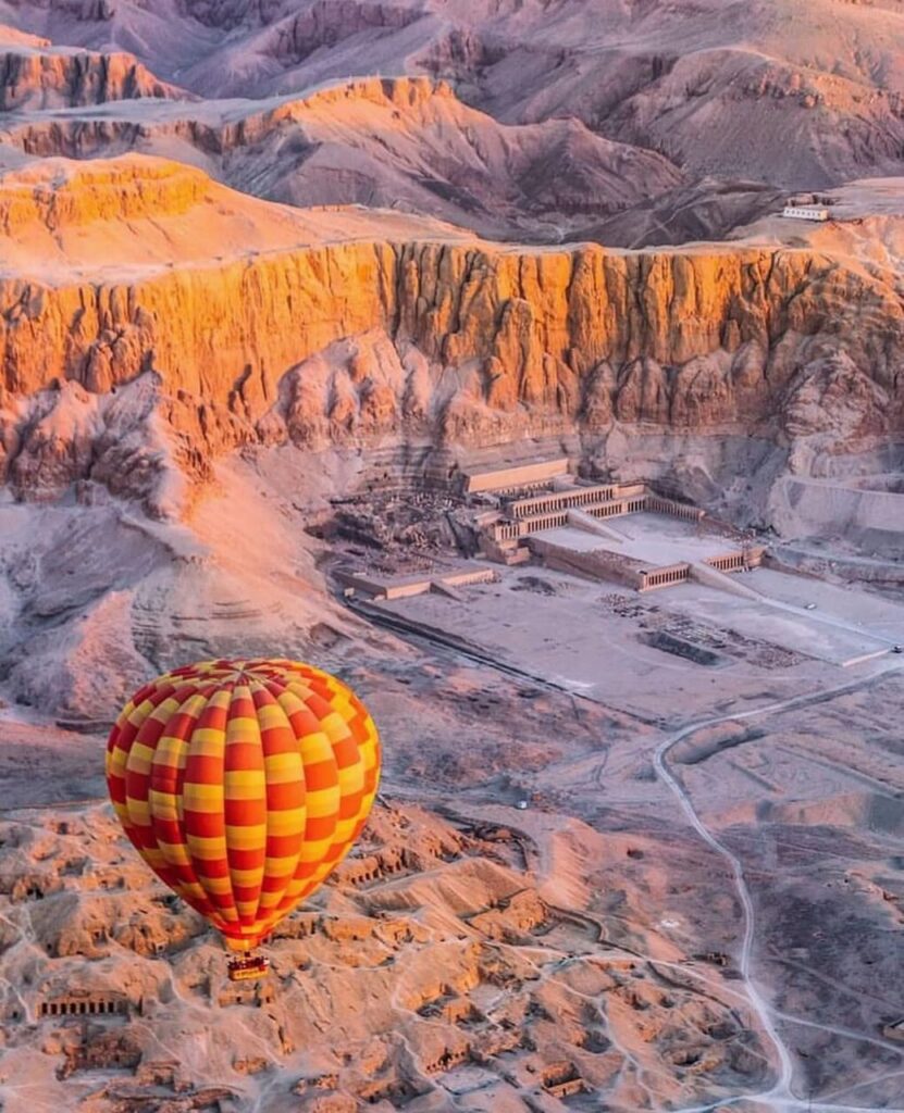 "Hot air balloon flying over the ancient sites of Luxor, Egypt, with the Temple of Hatshepsut and surrounding monuments visible at sunrise."